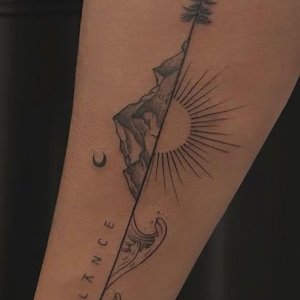 May include: A black ink tattoo on skin featuring a vertical line dividing two scenes. The top half shows a pine tree and mountain range next to a sun with rays. The bottom half depicts ocean waves and the word BALANCE written vertically.
