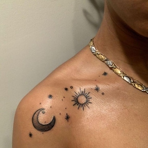 Peut inclure: Tatouage d'épaule représentant un croissant de lune, un soleil avec des lignes rayonnantes et plusieurs étoiles. Le tatouage est à l'encre noire et se trouve sur l'épaule d'une personne. Un collier en chaîne or et argent est également visible.