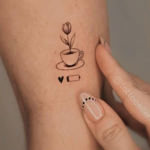 Peut inclure: Tatouage minimaliste représentant une tulipe poussant dans une tasse à thé sur une soucoupe. En dessous, un cœur et une icône de batterie. Le tatouage est à l'encre noire.