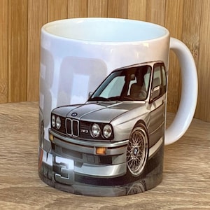 Könnte beinhalten: Weiße Keramik-Tasse mit einer silbernen Auto-Illustration. Das Auto-Design zeigt detaillierte Scheinwerfer, Räder und einen Kühlergrill. Auf der Tasse steht auch das Wort "M3".
