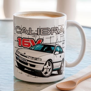 Pode incluir: Caneca de cerâmica branca com um gráfico de um carro branco e o texto "CALIBRA 16V". A caneca contém uma bebida clara. Uma chaleira de cobre e óculos de sol também são visíveis. A caneca tem cerca de 9 cm de altura.