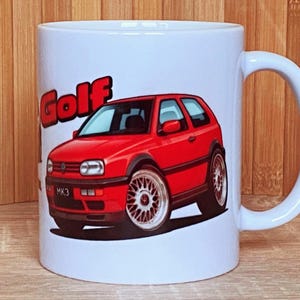 Puede incluir: Taza de cerámica blanca con un gráfico de coche rojo y la palabra "Golf" en letras rojas. El coche es un hatchback rojo con llantas plateadas. La taza tiene un asa blanca.
