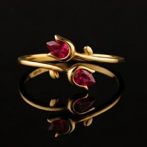 Puede incluir: Un anillo de oro con un diseño floral que presenta dos piedras preciosas rojas en forma de lágrima. El anillo está sobre un fondo negro, con su reflejo visible debajo. El diseño del anillo es delicado y elegante.