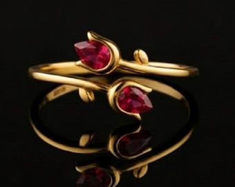 May include: A gold ring with a floral design featuring two red teardrop-shaped gemstones. The ring is set against a black background, with its reflection visible below. The ring's design is delicate and elegant.