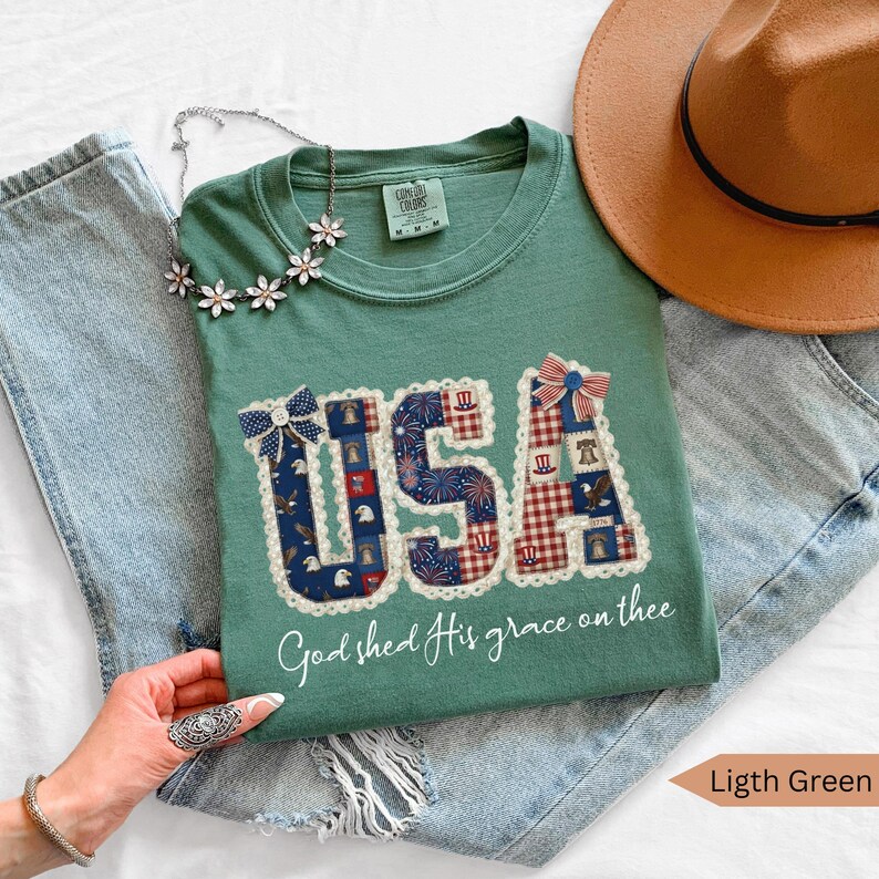 May include: Light green t-shirt with the word "USA" in a patchwork design featuring patriotic patterns. The shirt includes the phrase "God shed His grace on thee". A brown hat and a floral necklace are also visible.