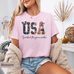 May include: A light pink t-shirt with the word "USA" in a patriotic design. The letters are filled with patterns of blue, red, and white. The text "God shed His grace on thee" is below the word "USA".