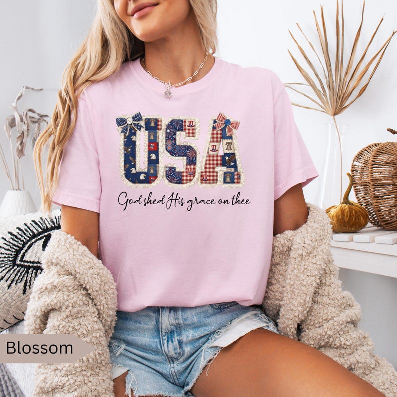 May include: A light pink t-shirt with the word "USA" in a patriotic design. The letters are filled with patterns of blue, red, and white. The text "God shed His grace on thee" is below the word "USA".