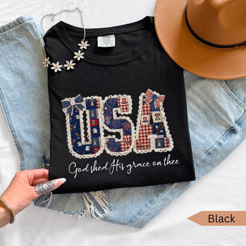 May include: Black t-shirt with a patriotic "USA" design in red, white, and blue, featuring fireworks, stars, and a gingham pattern. The shirt includes the phrase "God shed His grace on thee."