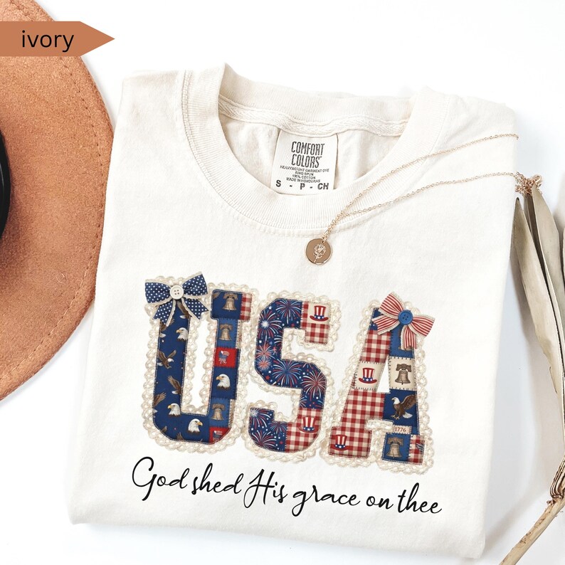 May include: Ivory-colored t-shirt with the word "USA" in a patchwork design featuring patriotic patterns. The shirt includes the phrase "God shed His grace on thee" and is accessorized with a gold necklace.
