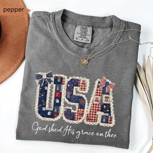 May include: Gray Comfort Colors t-shirt with a patriotic "USA" design in red, white, and blue patchwork. The shirt features a decorative bow and the text "God shed His grace on thee". A silver necklace with a pendant is draped on the shirt.