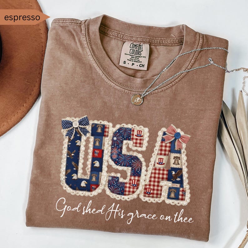 May include: Espresso-colored t-shirt with the word "USA" in a patchwork design featuring patriotic patterns and a scalloped edge. The shirt includes the phrase "God shed His grace on thee" in script. A silver necklace with a pendant is draped across the shirt.