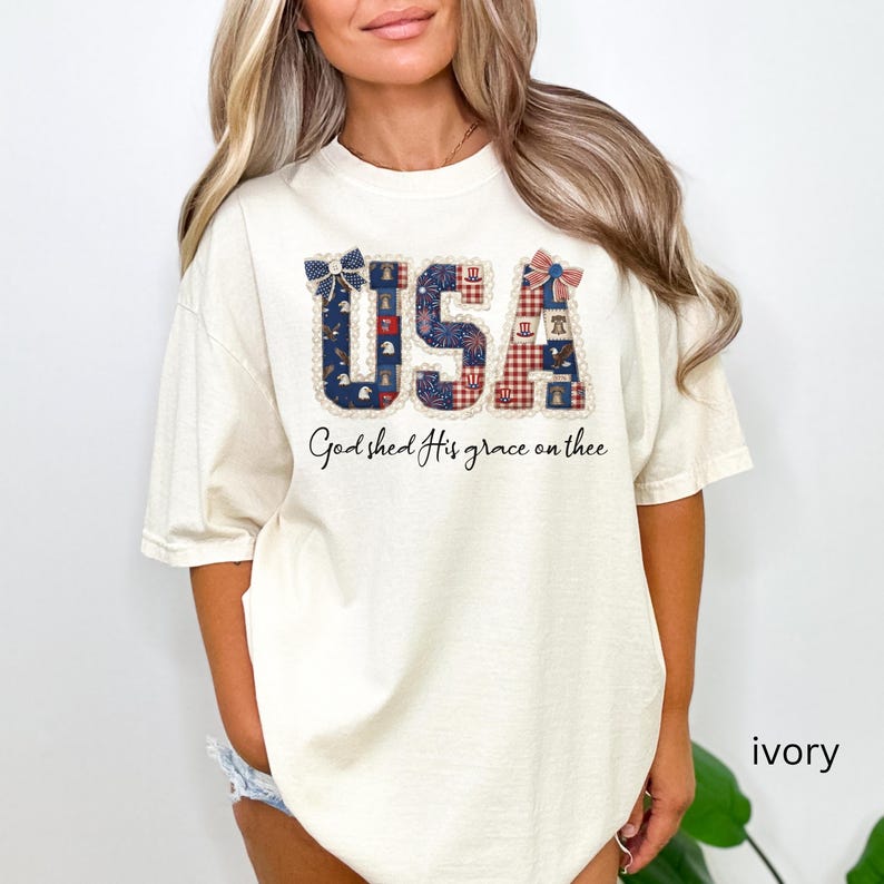 May include: Ivory-colored t-shirt with the word "USA" in a patriotic design. The letters are decorated with blue, red, and white patterns. Below the word "USA" is the phrase "God shed His grace on thee."
