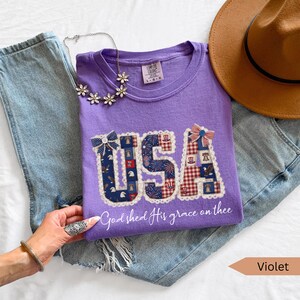 May include: A violet-colored t-shirt with the word "USA" in a patchwork design, featuring patriotic patterns. The shirt is paired with blue jeans, a brown hat, and a floral necklace. The text "God shed His grace on thee" is below the word "USA".