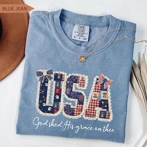 Flat lay of a Comfort Colors Blue Jean t-shirt featuring a detailed "USA" graphic in a coquette patchwork style. The letters are filled with vintage American patterns (eagles, liberty bells, fireworks) and adorned with faux-lace edges, a blue star bow, and a red striped bow. Below the graphic, the phrase "God shed His grace on thee" is written in elegant white script. The shirt is displayed with a brown hat, gold necklace, and dried palm leaves.