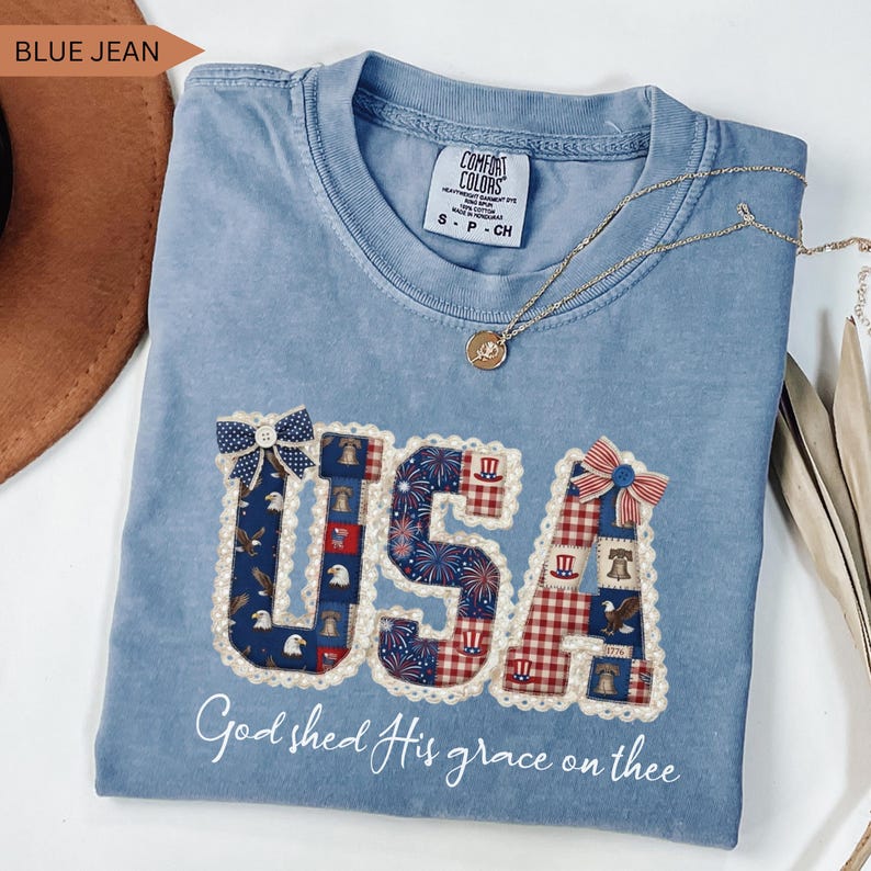 Flat lay of a Comfort Colors Blue Jean t-shirt featuring a detailed "USA" graphic in a coquette patchwork style. The letters are filled with vintage American patterns (eagles, liberty bells, fireworks) and adorned with faux-lace edges, a blue star bow, and a red striped bow. Below the graphic, the phrase "God shed His grace on thee" is written in elegant white script. The shirt is displayed with a brown hat, gold necklace, and dried palm leaves.