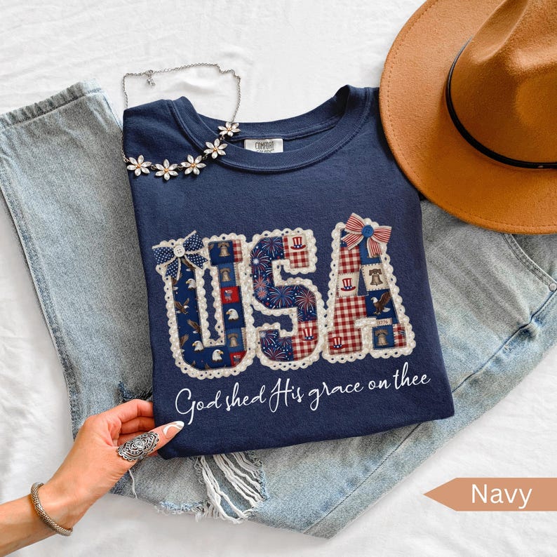 May include: Navy blue sweatshirt with the word "USA" in a patchwork design, featuring red, white, and blue patterns. The text "God shed His grace on thee" is below the word "USA". A brown hat and a floral necklace are also visible.