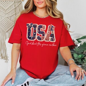 May include: A red t-shirt with the word "USA" in a patriotic design, featuring blue and red patterns and a white lace trim. The text "God shed His grace on thee" is printed below the graphic.