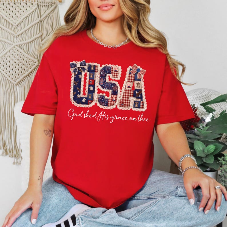 May include: A red t-shirt with the word "USA" in a patriotic design, featuring blue and red patterns and a white lace trim. The text "God shed His grace on thee" is printed below the graphic.
