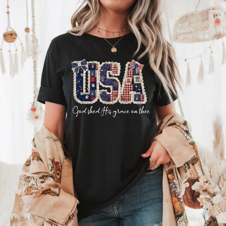 May include: Black t-shirt featuring the word "USA" in a patriotic design with red, white, and blue patterns. The text "God shed His grace on thee" is below the word "USA". The shirt has rolled-up sleeves.