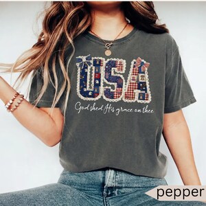 May include: Dark gray t-shirt with the word "USA" in a patchwork design featuring red, white, and blue patterns. The text "God shed His grace on thee" is written below the word "USA".