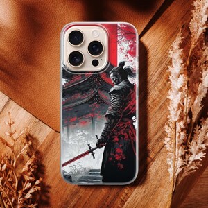 May include: A smartphone case featuring a Japanese samurai warrior with a red and black color scheme. The design includes a traditional building and a warrior holding a sword. The case is on a brown surface.