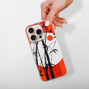 May include: A phone case featuring a Japanese-inspired design. The case has a white background with black bamboo stalks and leaves, a red sun, and a red border. The phone case is held by a hand.