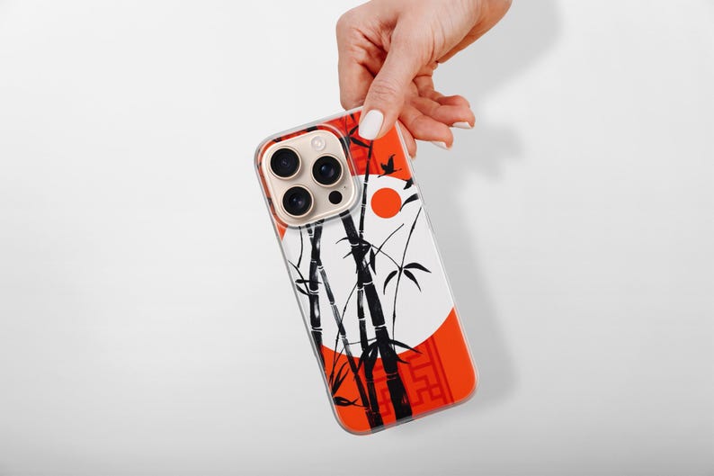 May include: A phone case featuring a Japanese-inspired design. The case has a white background with black bamboo stalks and leaves, a red sun, and a red border. The phone case is held by a hand.