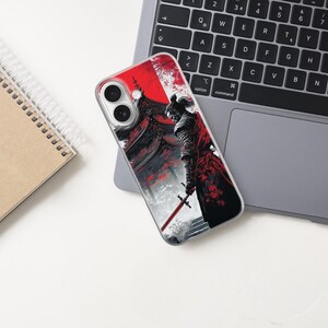 May include: A smartphone case featuring a detailed illustration of a samurai warrior with a red and black color scheme. The design includes a traditional Japanese building and a sword. The case is on a laptop keyboard and a notebook.