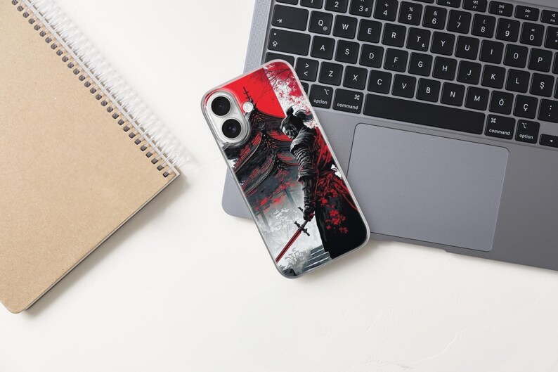 May include: A smartphone case featuring a detailed illustration of a samurai warrior with a red and black color scheme. The design includes a traditional Japanese building and a sword. The case is on a laptop keyboard and a notebook.