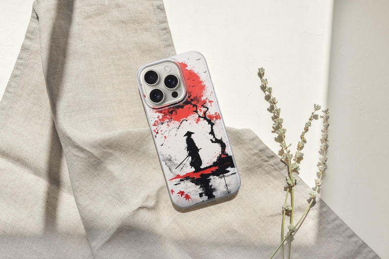 May include: A white phone case featuring a Japanese-style illustration of a samurai warrior standing in front of a red flowering tree. The design is in black, red, and white, with a minimalist aesthetic. The case has a clear border and a camera cutout.