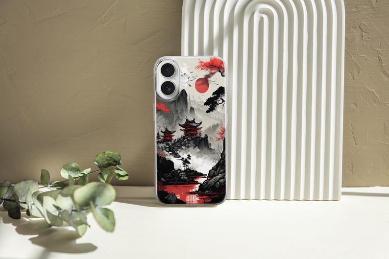May include: A clear phone case featuring a Japanese landscape design. The artwork depicts red pagodas, a red sun, and a river, set against a backdrop of mountains and trees in shades of black, gray, and red.