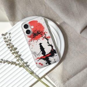 May include: A phone case featuring a Japanese-inspired design. The case displays a red sun, a silhouetted samurai, and a tree with red leaves. The design is set against a white background with black and gray accents.