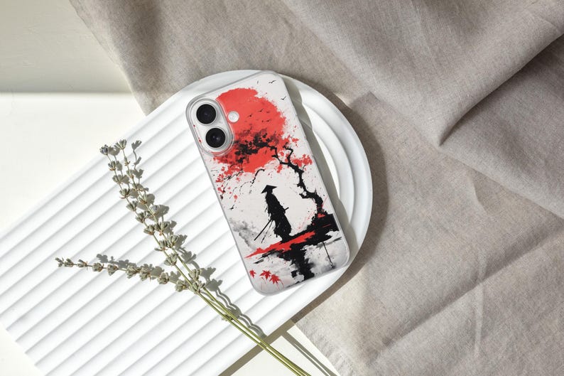 May include: A phone case featuring a Japanese-inspired design. The case displays a red sun, a silhouetted samurai, and a tree with red leaves. The design is set against a white background with black and gray accents.
