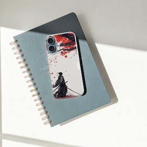 May include: A clear phone case featuring a Japanese samurai warrior design with red and white accents. The phone case is resting on a blue spiral notebook with gold-colored binding.