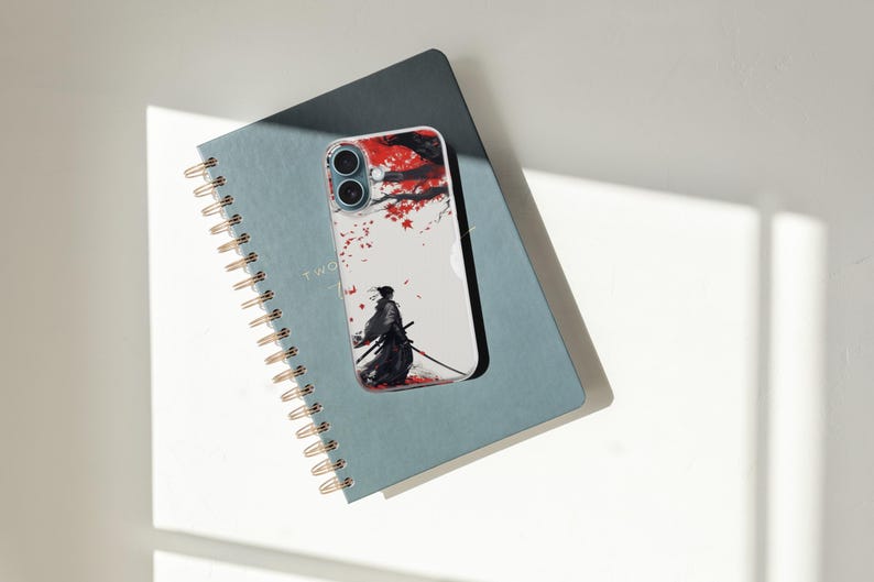 May include: A clear phone case featuring a Japanese samurai warrior design with red and white accents. The phone case is resting on a blue spiral notebook with gold-colored binding.