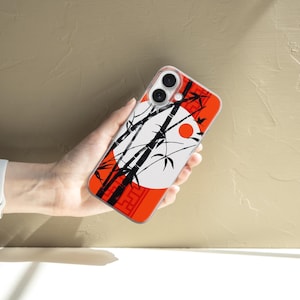 May include: A phone case featuring a Japanese-inspired design. The case has a red border with a white circle and a red sun. Black bamboo stalks and leaves are silhouetted against the white circle. The phone case is held in a hand.