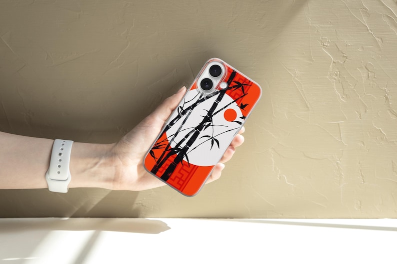 May include: A phone case featuring a Japanese-inspired design. The case has a red border with a white circle and a red sun. Black bamboo stalks and leaves are silhouetted against the white circle. The phone case is held in a hand.