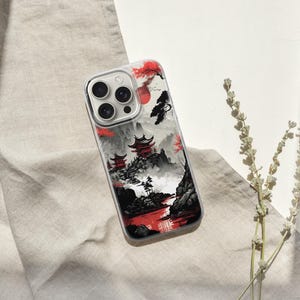 May include: A clear phone case featuring a Japanese landscape design with red accents. The case showcases a mountain scene with pagodas, a red sun, and a river. The case is resting on a beige fabric.