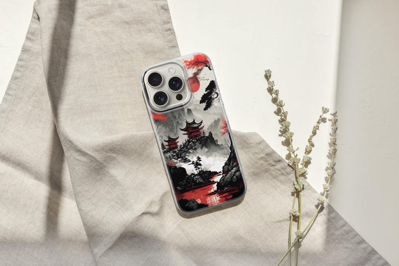 May include: A clear phone case featuring a Japanese landscape design with red accents. The case showcases a mountain scene with pagodas, a red sun, and a river. The case is resting on a beige fabric.