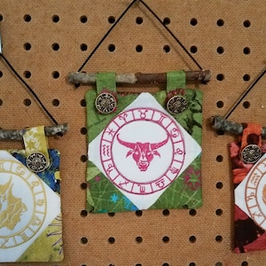 May include: Three fabric wall hangings with a stick at the top for hanging. Each hanging features a different zodiac sign: Taurus, Gemini, and Aries. The signs are embroidered in white on a white background with a circle of zodiac symbols around the outside. The hangings are made of different fabrics with a patchwork design.