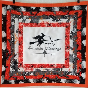 May include: A white fabric square with a black embroidered image of a witch flying on a broomstick with bats. The fabric is surrounded by layers of orange, black, and gray fabric with a spiderweb pattern. The text "Samhain Blessings" is embroidered below the witch.