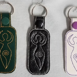 May include: Three leather keychains with a stylized female figure design. The keychains are green, black, and white with gold, silver, and purple accents.