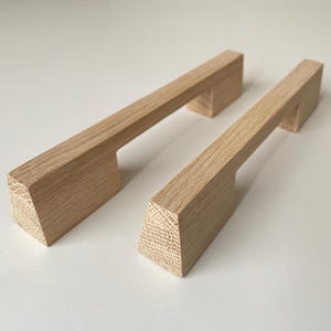 Op de afbeelding: Twee lichtgekleurde houten kastgrepen met een natuurlijk nerfpatroon. Elke greep heeft een lange, rechthoekige bovenbalk ondersteund door twee schuine, trapeziumvormige voetjes aan beide uiteinden, met een gladde, onafgewerkte houttextuur.