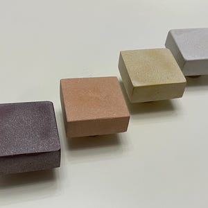Può includere: Quattro pomelli quadrati per mobili in vari colori: viola scuro, marrone rossiccio, beige e grigio chiaro. Ogni pomello ha una parte superiore piatta e un bordo leggermente smussato. I pomelli sono disposti diagonalmente su una superficie bianca.