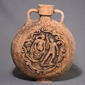 May include: A terracotta-colored, round flask with two handles and a small spout. The flask features a raised relief of two figures within a decorative border. The surface has a textured, speckled appearance.