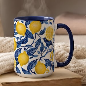 May include: A ceramic mug with a vibrant lemon and leaf pattern. The mug features a white background with yellow lemons and blue leaves. The interior and handle are a deep blue color. The mug is sitting on a book.