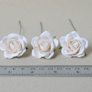 May include: Three ivory paper roses with green stems, each measuring approximately 1.5 inches in diameter, are arranged on a white surface with a ruler for scale.