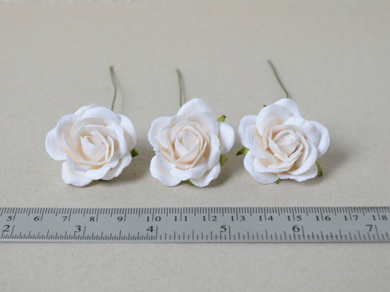 May include: Three ivory paper roses with green stems, each measuring approximately 1.5 inches in diameter, are arranged on a white surface with a ruler for scale.