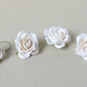 May include: Four white paper roses with green stems. The roses are arranged in a row on a light brown surface.