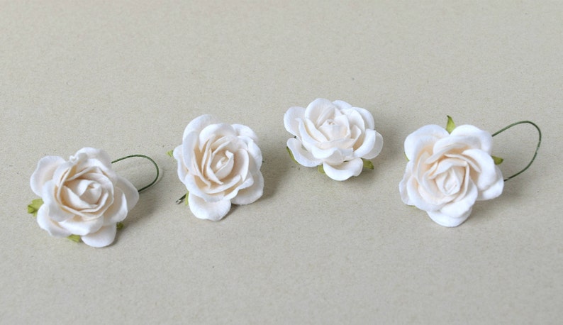 May include: Four white paper roses with green stems. The roses are arranged in a row on a light brown surface.